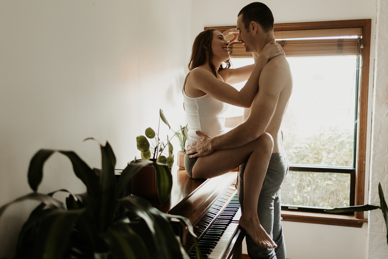 woman sits on piano with plants around her while man stands between her legs holding her