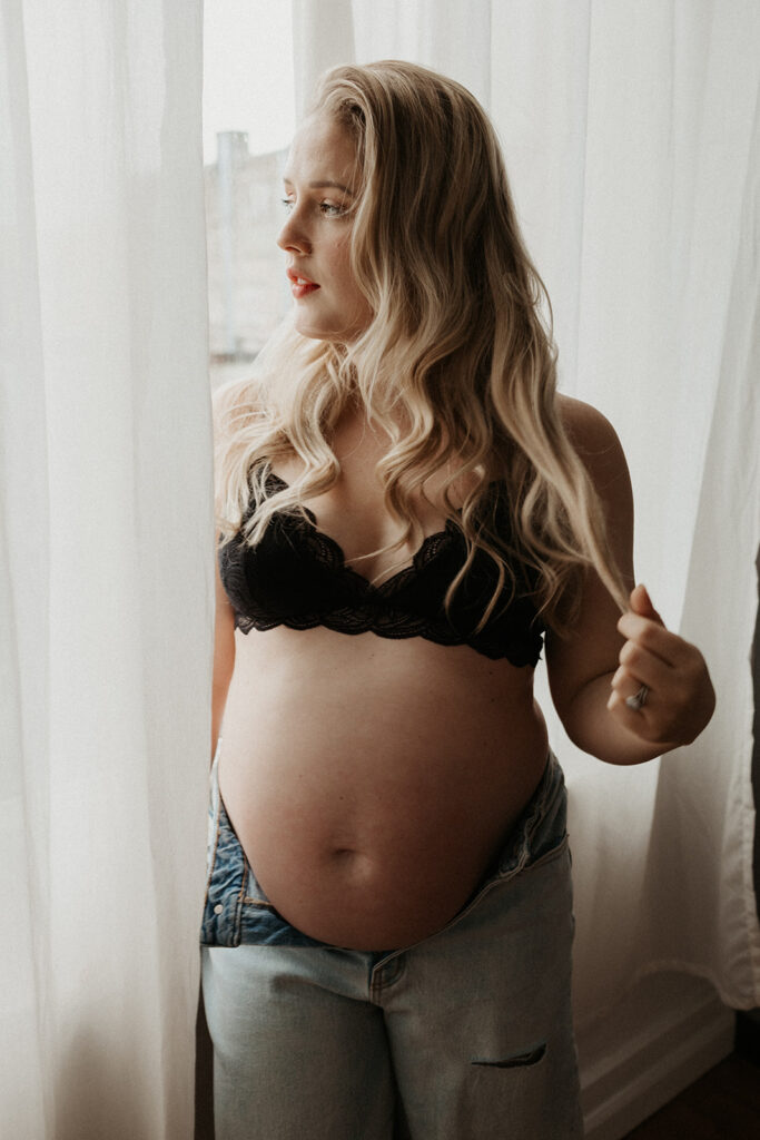 pregnant woman wears jeans and bra, sits on window sill while playing with hair
