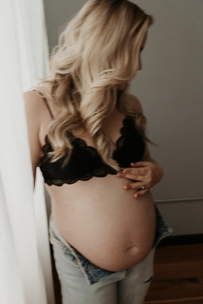 pregnant white woman plays with hair while standing by curtains