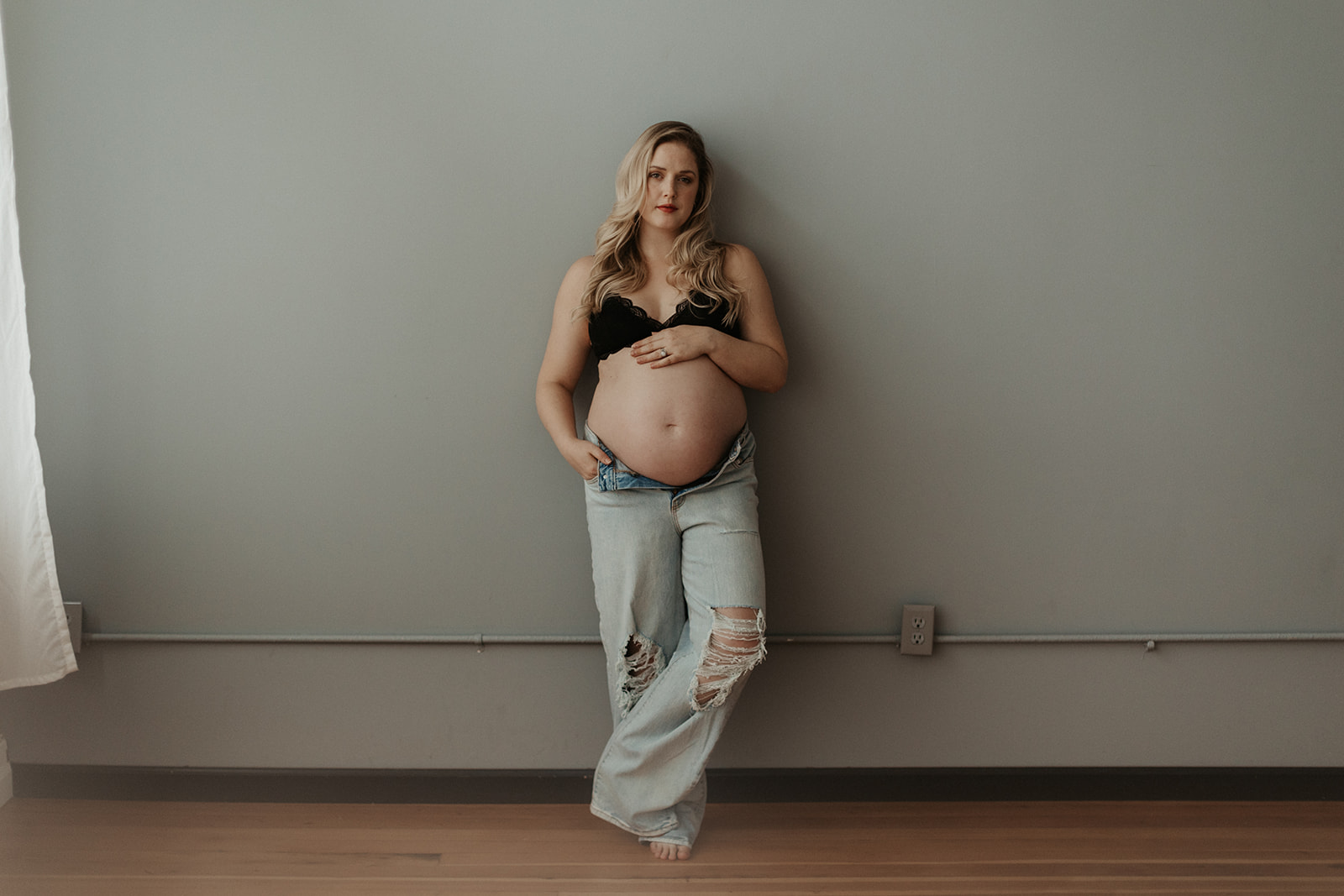 maternity boudoir, white woman in baggy jeans and bra standing against wall