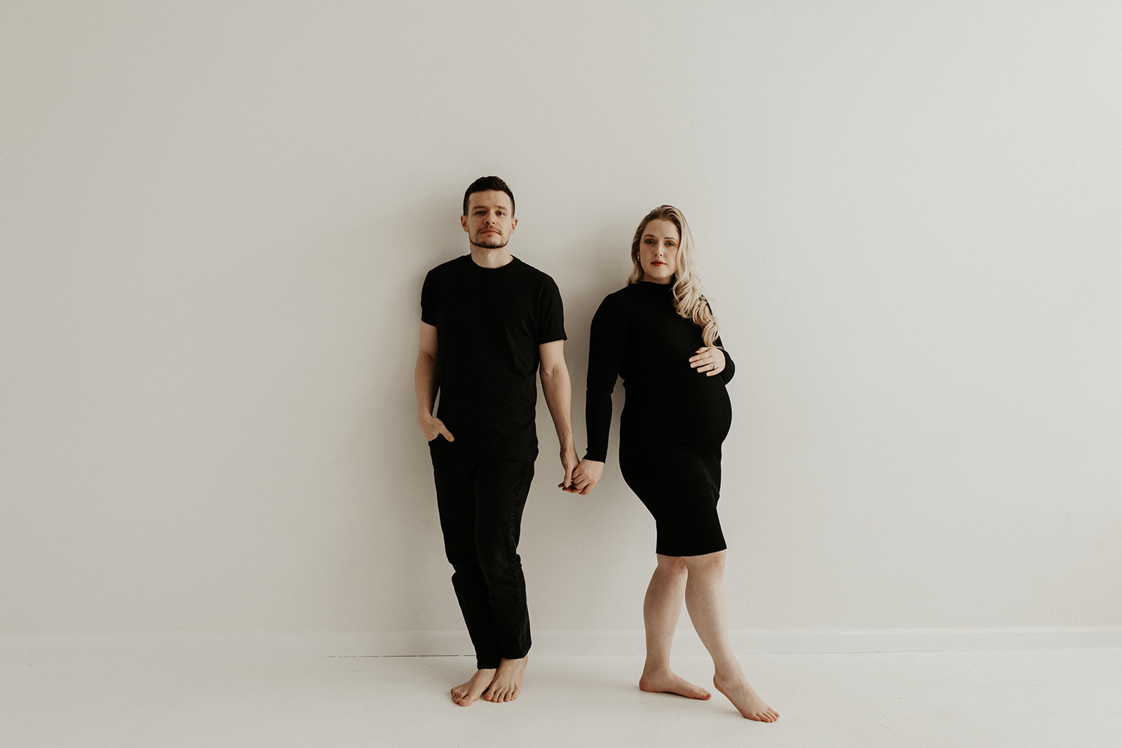 white couple wearing all black stand holding hands against white wall
