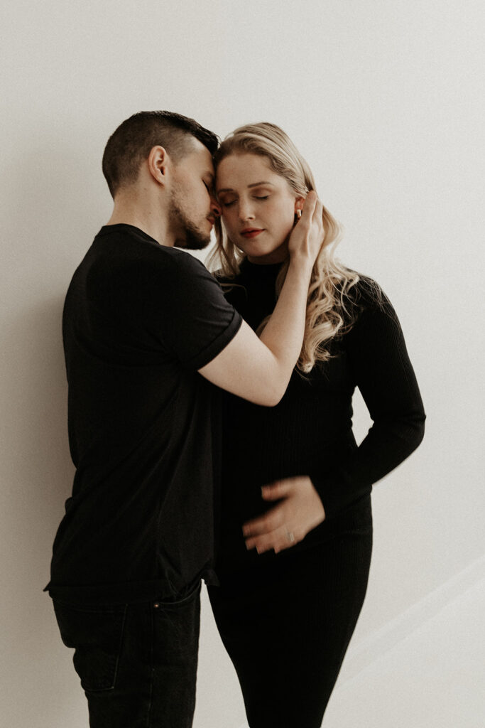 pregnant woman holds stomach while man cradles her face and leans in to nuzzle her