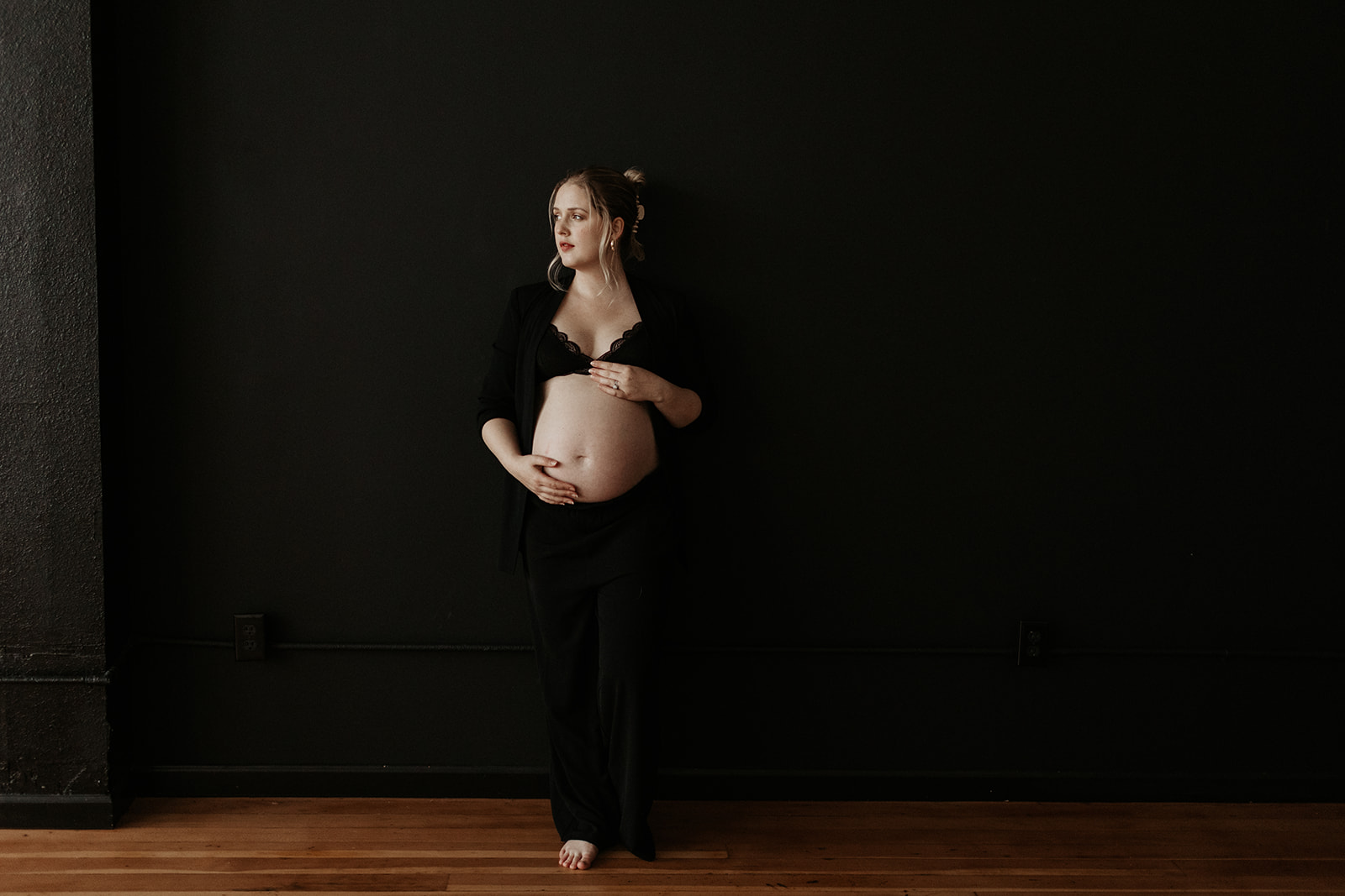 maternity boudoir, pregnant woman wears black pants and black bra against black backdrop