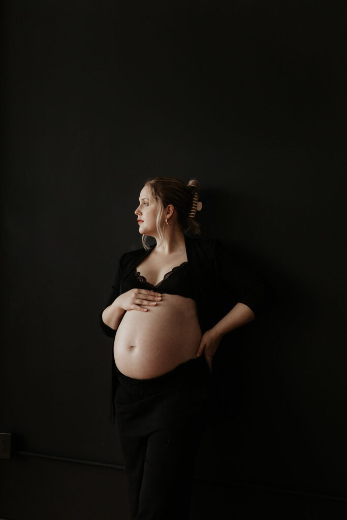 maternity boudoir, pregnant woman wears black pants and black bra against black backdrop