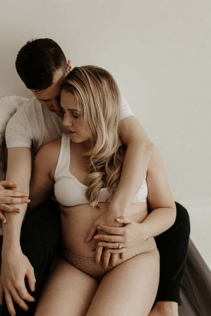 couples boudoir, pregnant woman sits in between her husband legs as he hugs her from behind