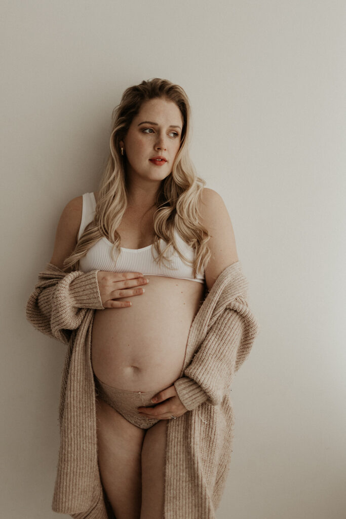 maternity boudoir, pregnant woman wearing nude underwear and white bra stands against white wall holding stomach