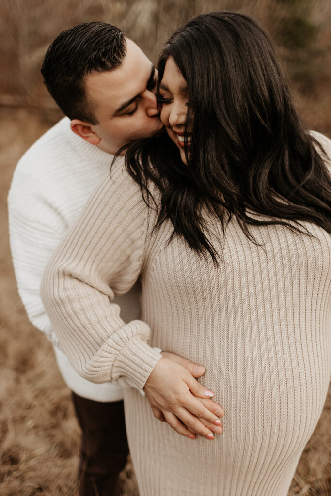 man kisses pregnant woman's cheek from behind holding her stomach during maternity shoot