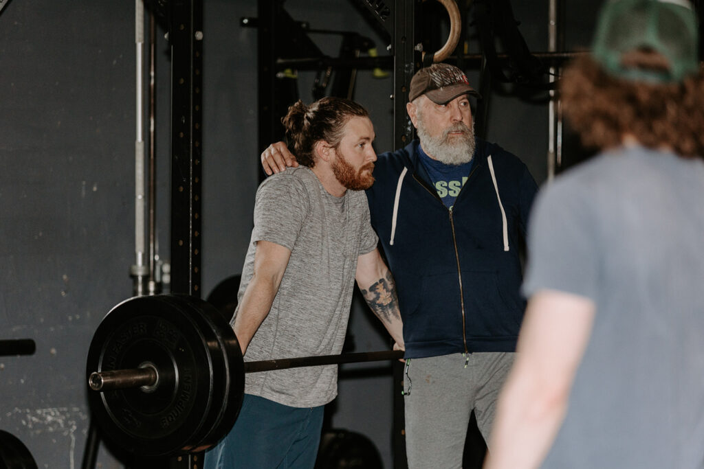 older man touching a younger man on the shoulder while they lean over weights