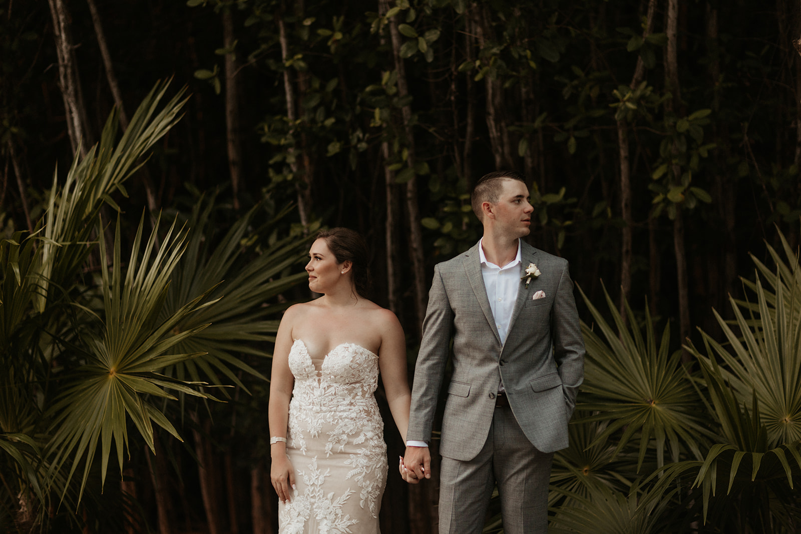 bride and groom stand holding hands, looking in opposite directions against a forest of tropical plants